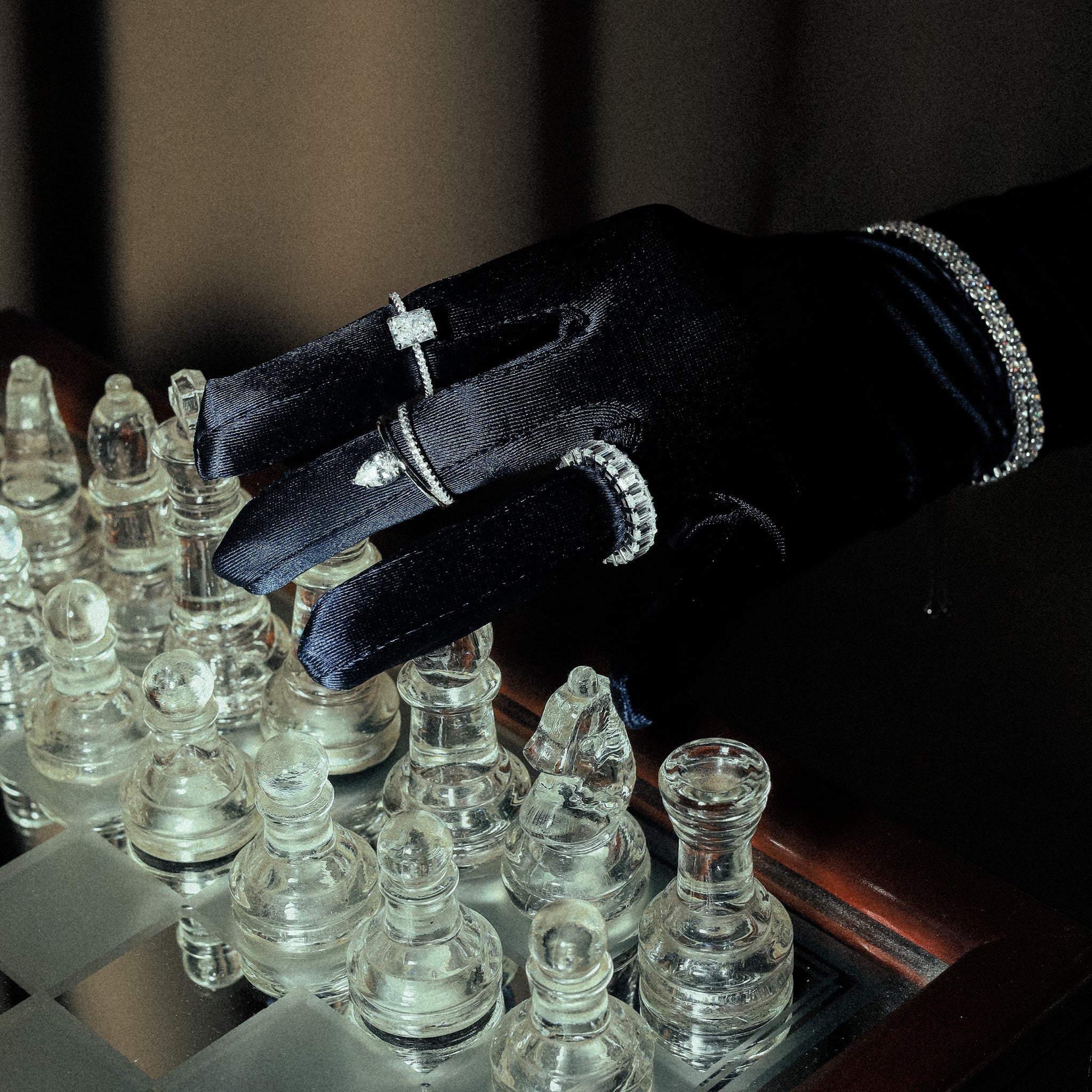 Kapi Rectangular Cut Ring in Silver worn on finger, emphasizing the elegant design with rectangular center stone and cubic zirconia accents, set in 925 sterling silver for a sophisticated look. Rhodium plated. Worn on a hand with a satin navy blue glove, featured in a stunning jewelry stack. The picture is set in an elegant surrounding featuring a classy chess set made of glass