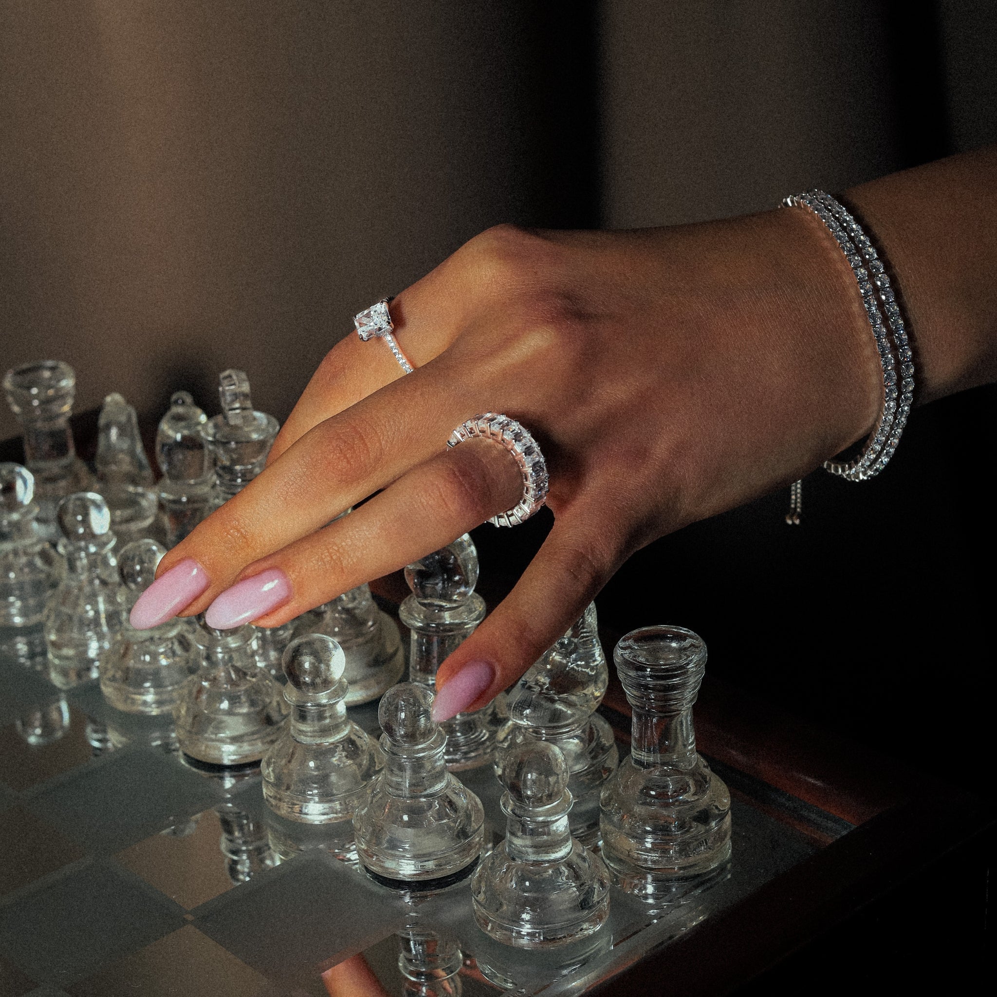 Kapi Rectangular Cut Ring in Silver worn on finger, emphasizing the elegant design with rectangular center stone and cubic zirconia accents, set in 925 sterling silver for a sophisticated look. Rhodium plated. Featured in a stunning image of a glass chess, and a hand stacked with tennis bracelets and beautiful rings.