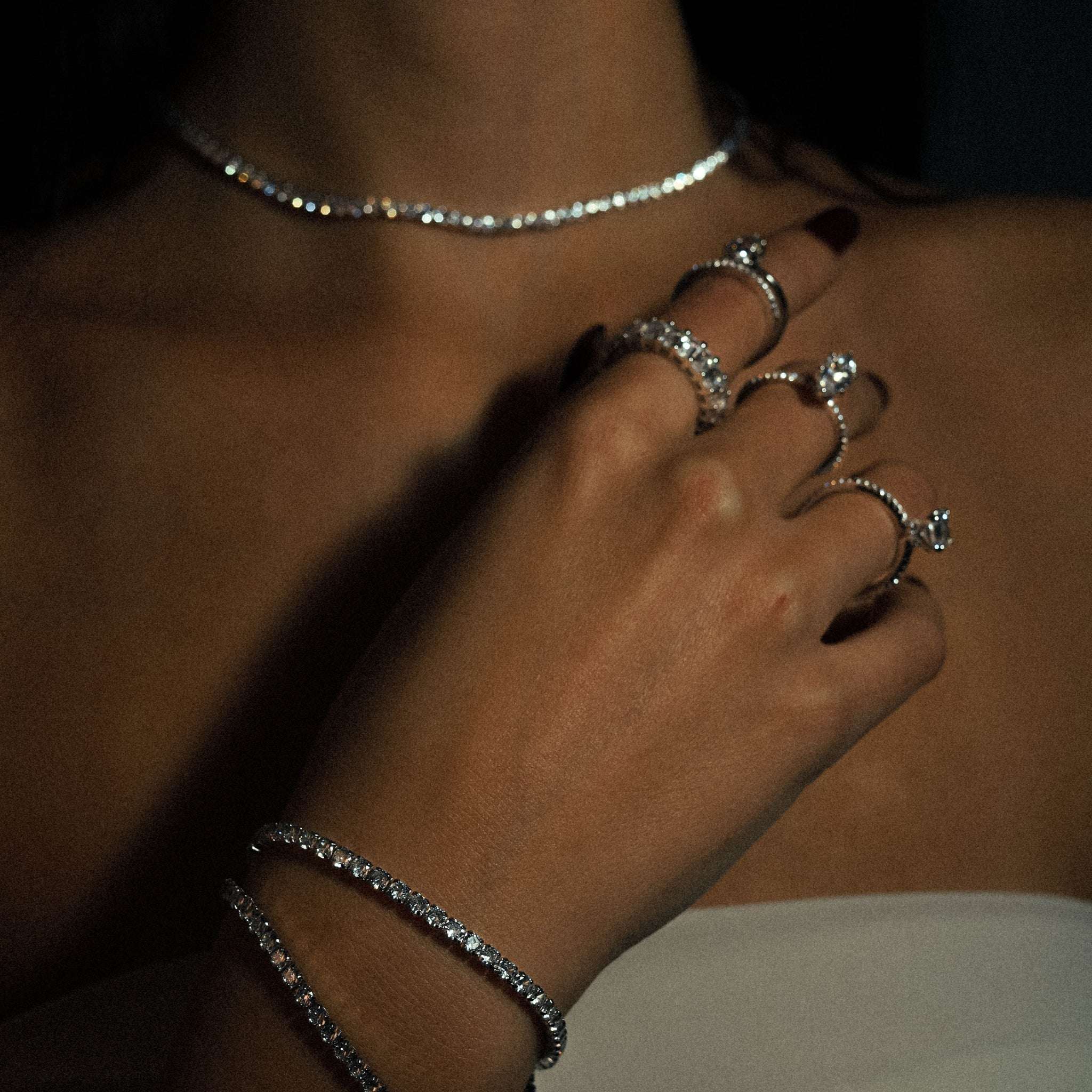 Silver Tennis Necklace worn around the neck, displaying the radiant cubic zirconia stones set in a minimalist sterling silver design, offering a timeless and sophisticated accessory. Rhodium plated. Featured in a stunning and luxurious picture, featuring multiple rings and tennis bracelets.
