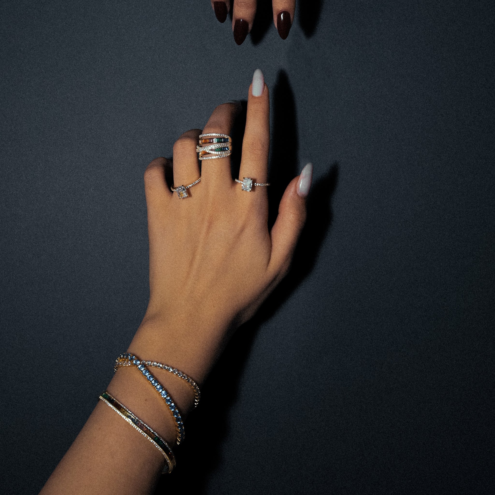 Aqua Dream Tennis Bracelet with gold plating and round blue cubic zirconia stones, featuring a secure clasp mechanism for enhanced safety. Worn on wrist. Chroma Bangle in Silver worn on wrist, showcasing its asymmetrical 925 sterling silver design with rhodium plating and colorful AAAAA grade cubic zirconia accents. Chroma Ring in gold-plated sterling silver worn on finger, displaying its eye-catching asymmetrical design and colorful AAAAA grade cubic zirconia stones for a luxurious, fashionable app